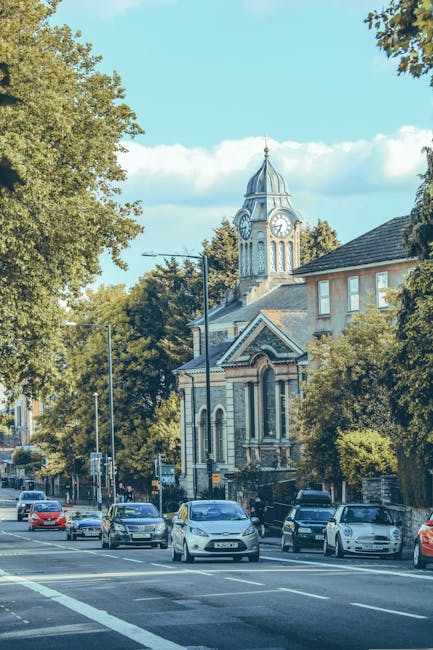 A street scene in Balham with several parked and moving cars along the roadside, facing toward a historic church building with a large clock tower and a pointed spire, situated amidst trees and reside