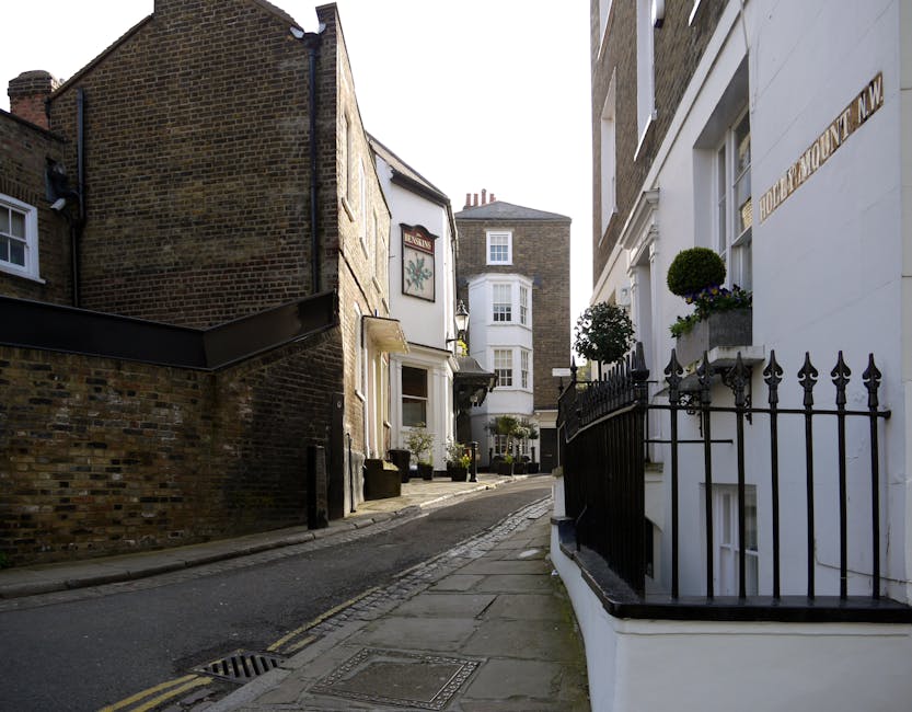 A street scene in Balham with several parked and moving cars along the roadside, facing toward a historic church building with a large clock tower and a pointed spire, situated amidst trees and reside